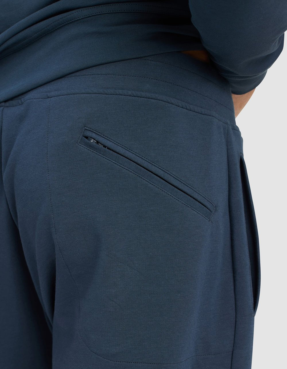 Close-up of a person wearing Cozy Earth Men's StretchTech Quarter Zip & Jogger Set in dark blue, showing a zippered back pocket and one hand partly in the waistband; plain light gray background.|Color:Eclipse