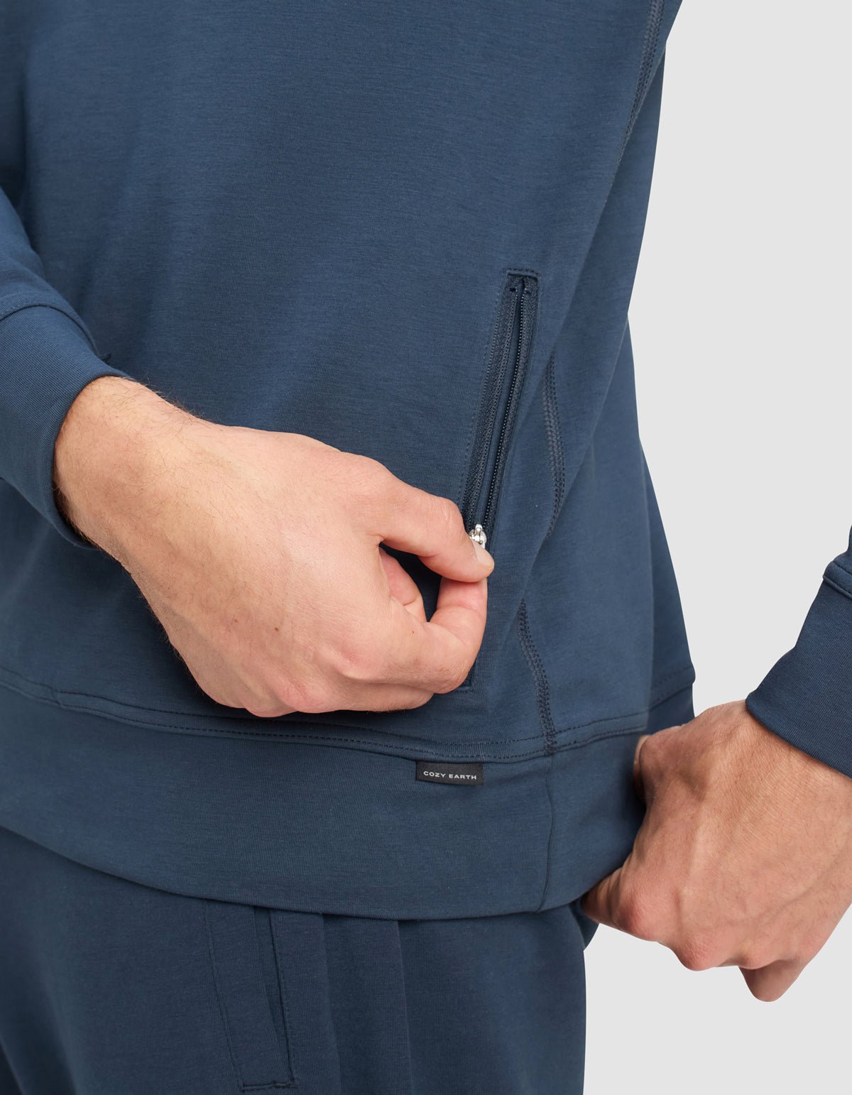A man wearing Cozy Earth Men's StretchTech Quarter Zip and matching pants is pulling the zipper of a side pocket on the top, set against a plain light gray background.|Color:Eclipse