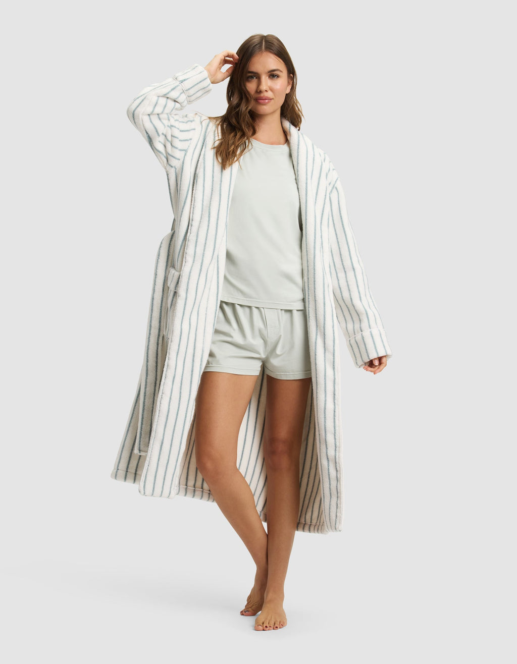 A woman stands barefoot, smiling with one hand in her hair, wearing the Cozy Earth Striped Luxe Bath Robe—a long, white robe with thin blue stripes—over a light-colored pajama set with shorts. |Color:Brook Stripe