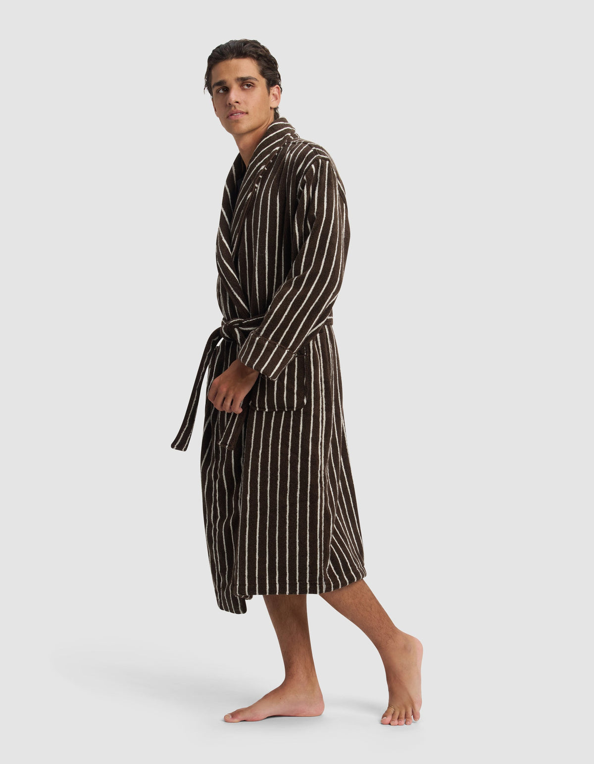 A young man stands barefoot against a white background, wearing Cozy Earth's Striped Luxe Bath Robe in dark brown with vertical white stripes and a waist tie, gazing slightly to the side.|Color:Espresso Coconut Stripe
