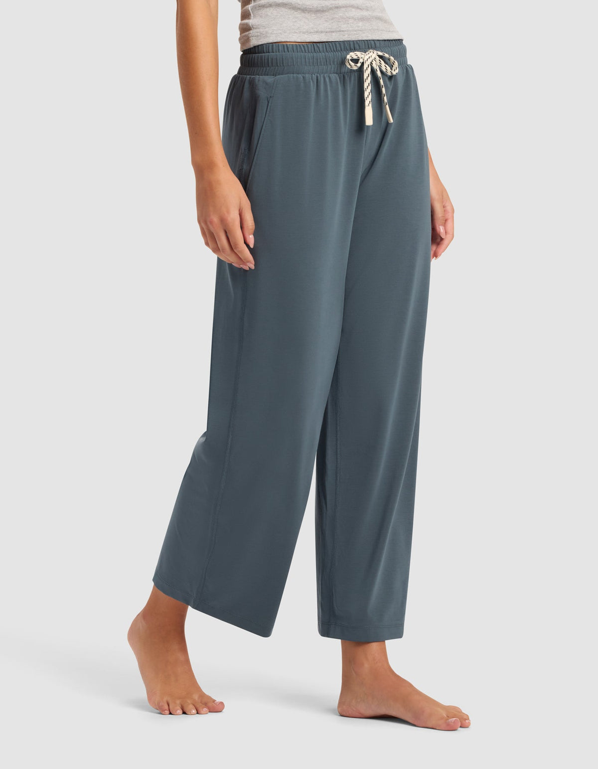 A woman wears Cozy Earth's Women's Studio Cropped Wide Leg Pant—loose, blue-gray with an elastic waistband and cream drawstring—standing barefoot against a plain light background.|Color:Dawn
