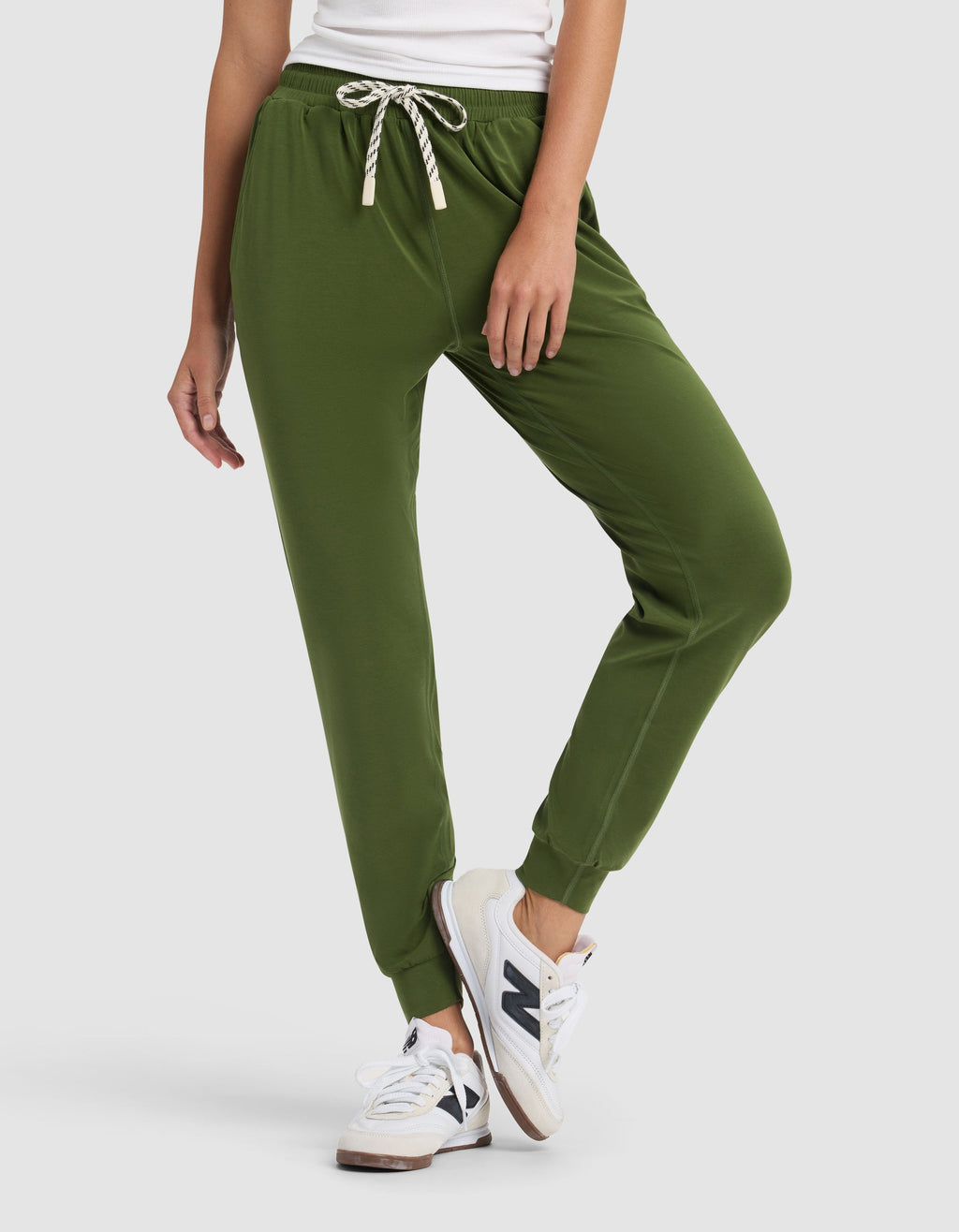 A woman stands against a plain light background wearing a white top, Cozy Earth Women's Studio Jogger pants in olive green with a drawstring, and white New Balance sneakers, one leg slightly bent.|Color:Grove