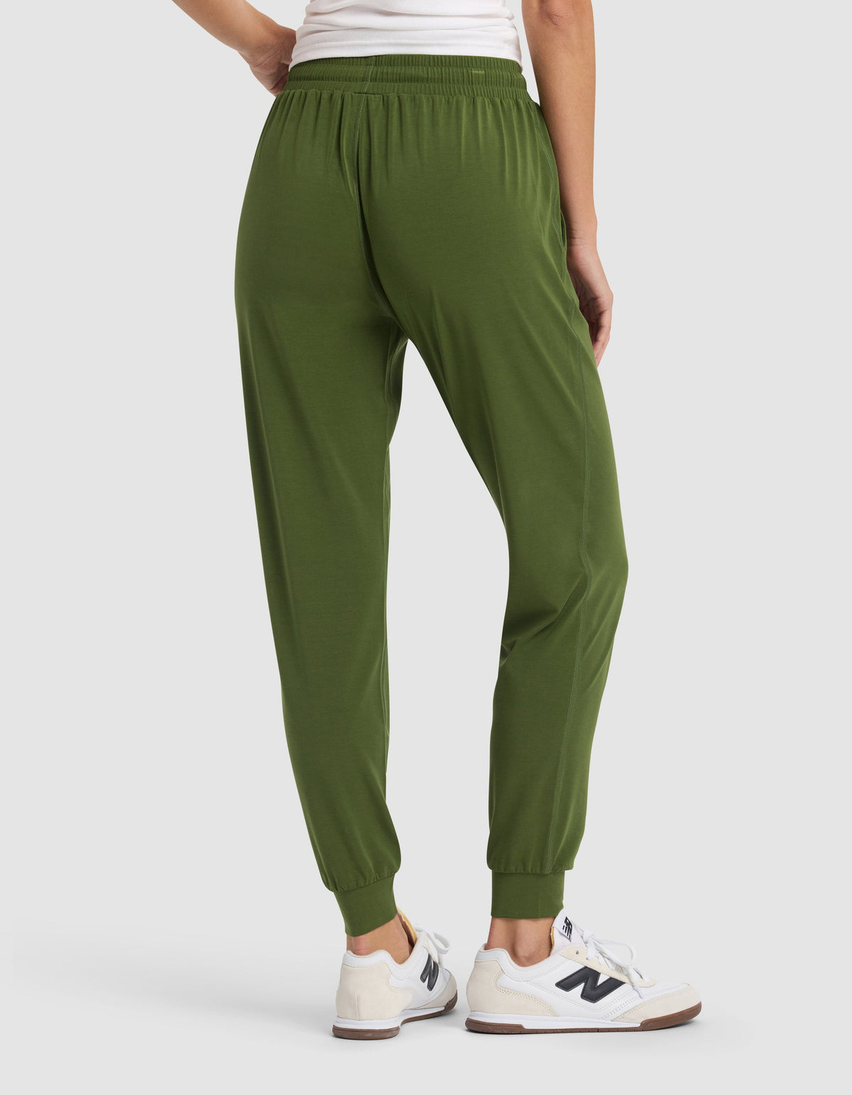 A woman is shown from the waist down wearing Cozy Earth Women's Studio Jogger pants in olive green, paired with white sneakers, standing on a light gray background.|Color:Grove
