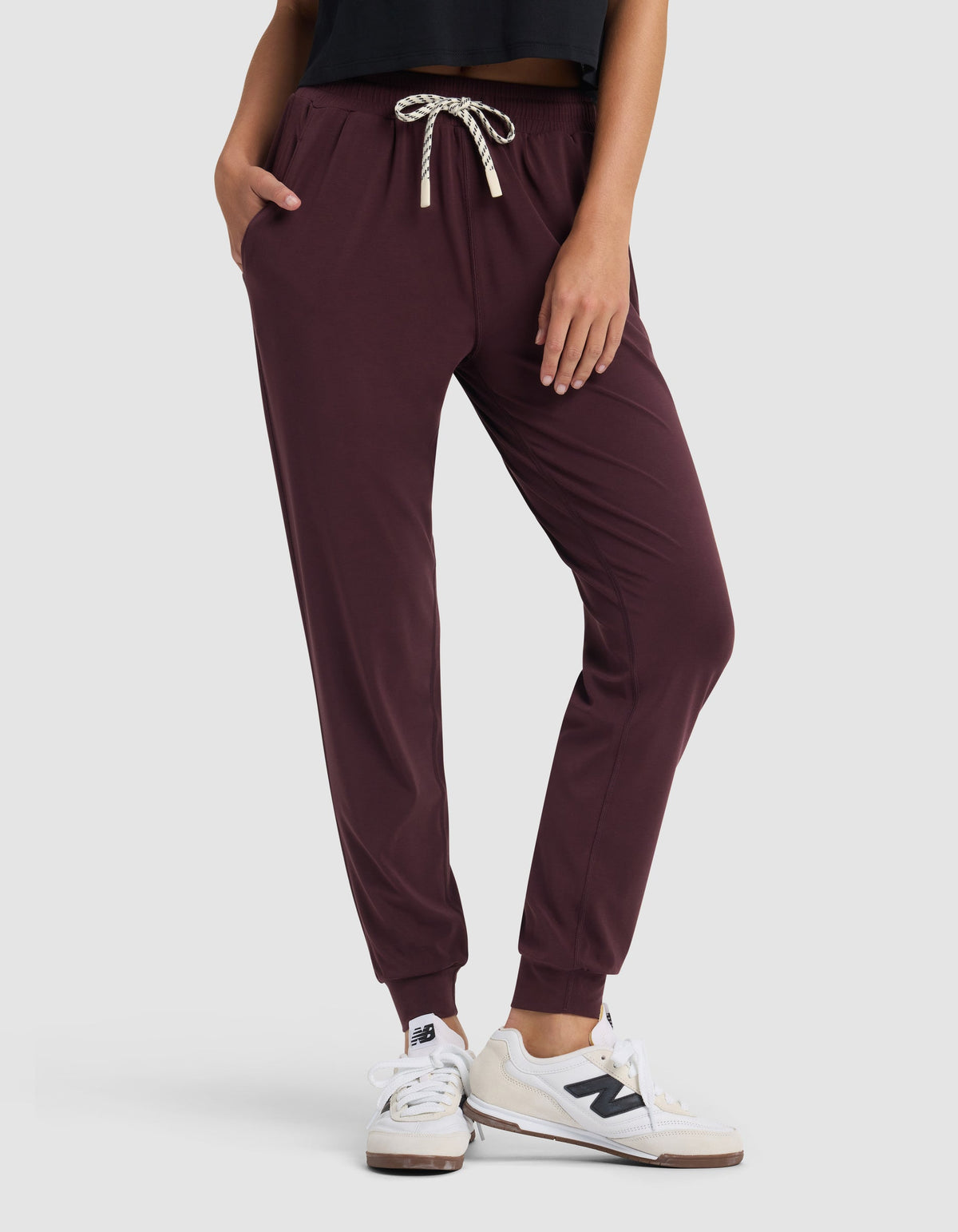 A person stands with hands in pockets against a plain light background, wearing Cozy Earth Women's Studio Jogger pants in maroon, a black top, and white New Balance sneakers.|Color:Raisin