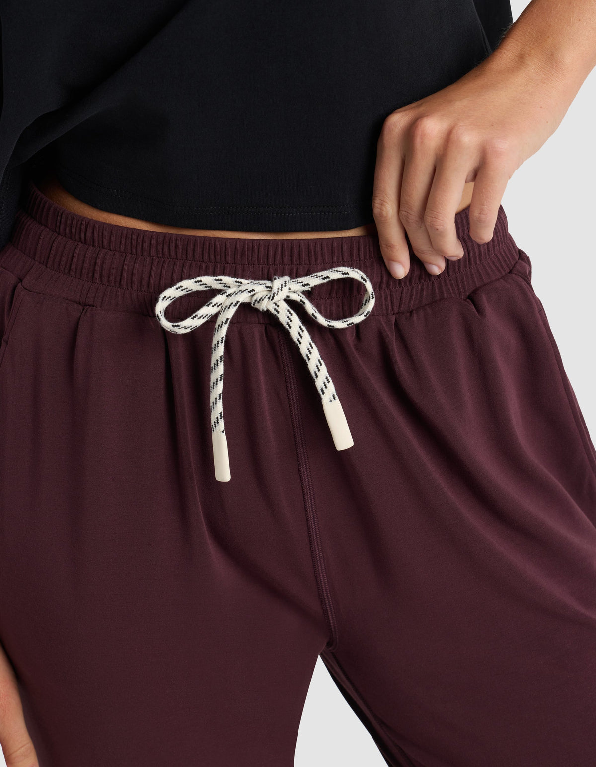 A woman models the Cozy Earth Women's Studio Jogger in maroon, featuring a white and black patterned drawstring tied in a bow, paired with a black top and posing with one hand on her hip.|Color:Raisin