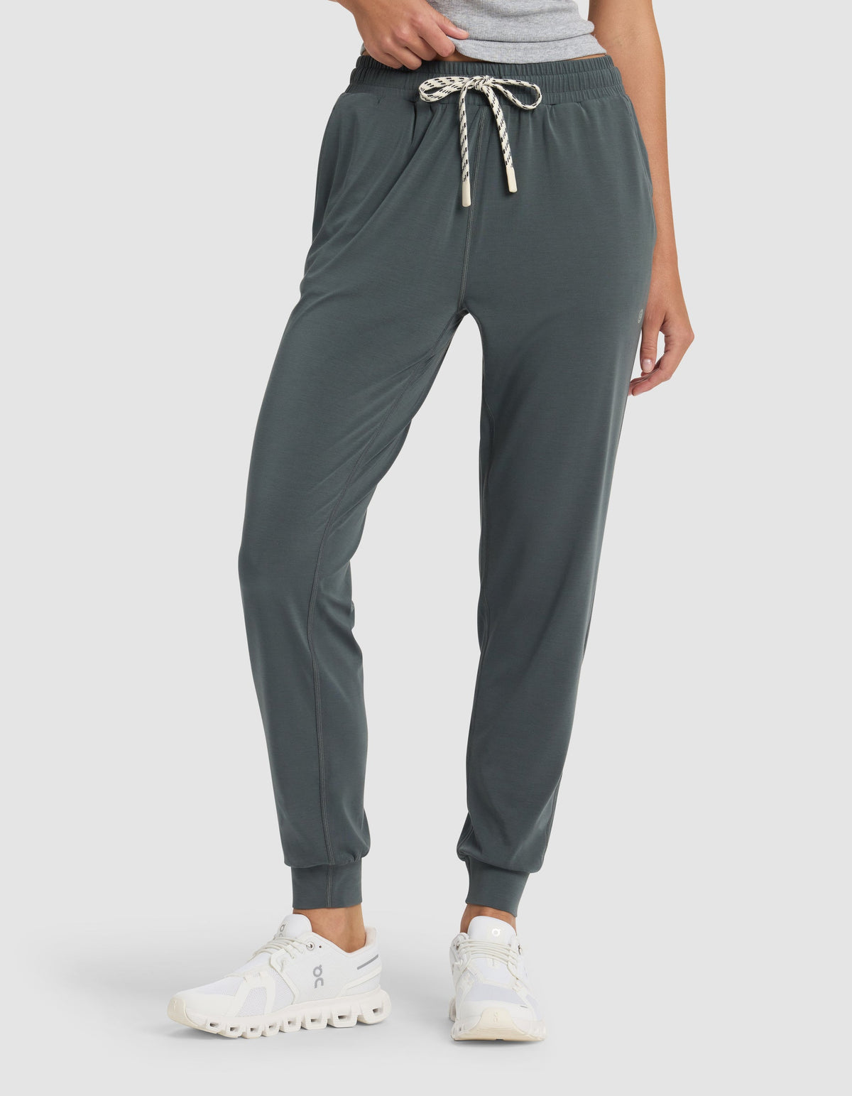 A person wears Cozy Earth's Women's Studio Jogger in dark green with a striped drawstring, white sneakers, and a gray top. The photo captures the outfit from the waist down against a plain background.|Color:Storm