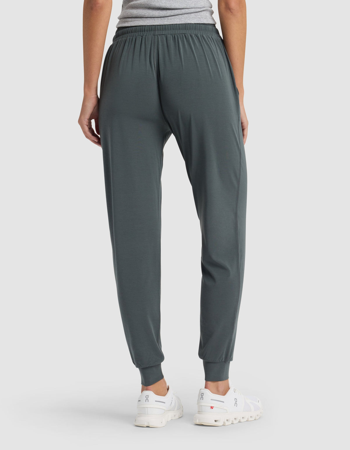 A person faces away, wearing Cozy Earth's Women's Studio Jogger in dark green, a grey shirt, and white athletic shoes against a plain light background.|Color:Storm
