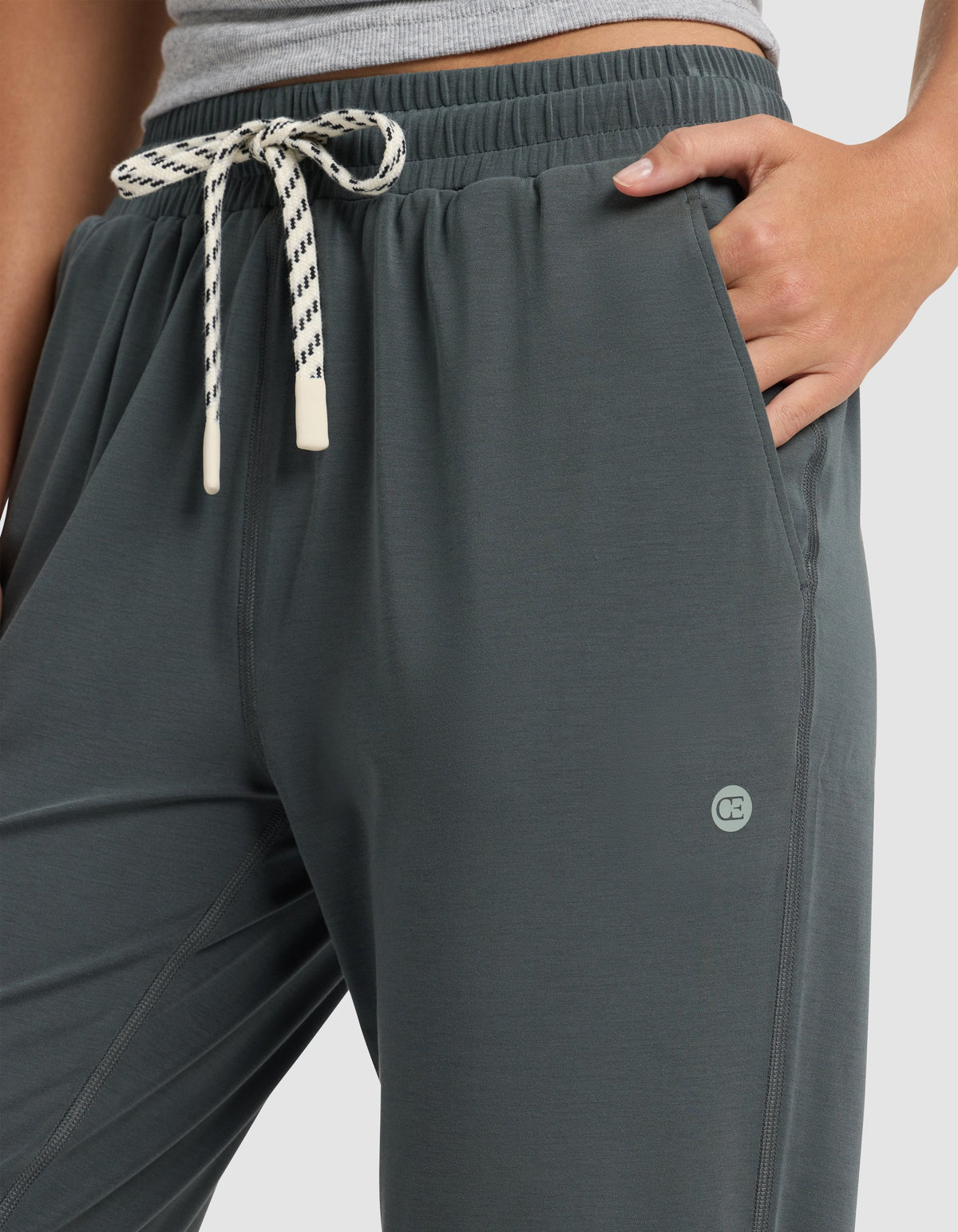 A person wears Cozy Earth's Women's Studio Jogger in dark grey, featuring a white and black drawstring, small circular logo by the left pocket, paired with a light grey top and one hand in the jogger's pocket.|Color:Storm
