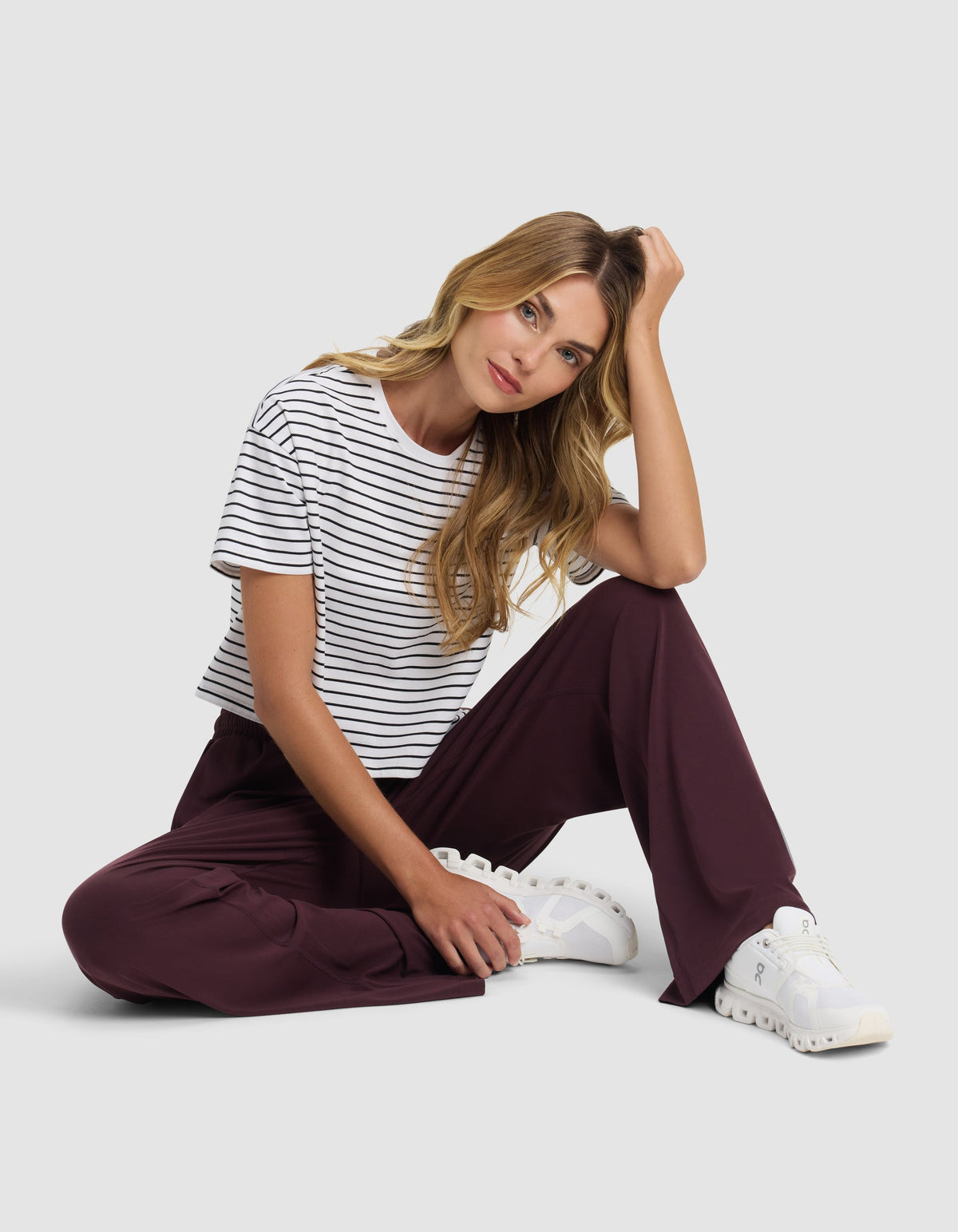 A woman with long blonde hair sits on the floor against a plain background, wearing Cozy Earth's Women's Studio Wide Leg Pant in maroon, paired with a black and white striped t-shirt and white sneakers. She rests her head on one hand, gazing at the camera.|Color:Raisin