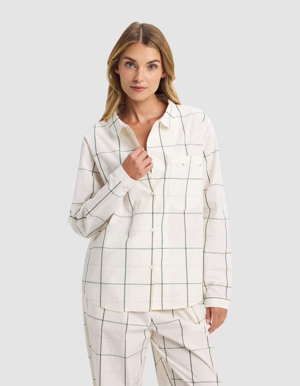 A woman stands against a plain background wearing the Cozy Earth Women's Sutton Long Sleeve Pajama Top in white with a green grid pattern, facing forward and touching her collar.|Color:Bone Windowpane