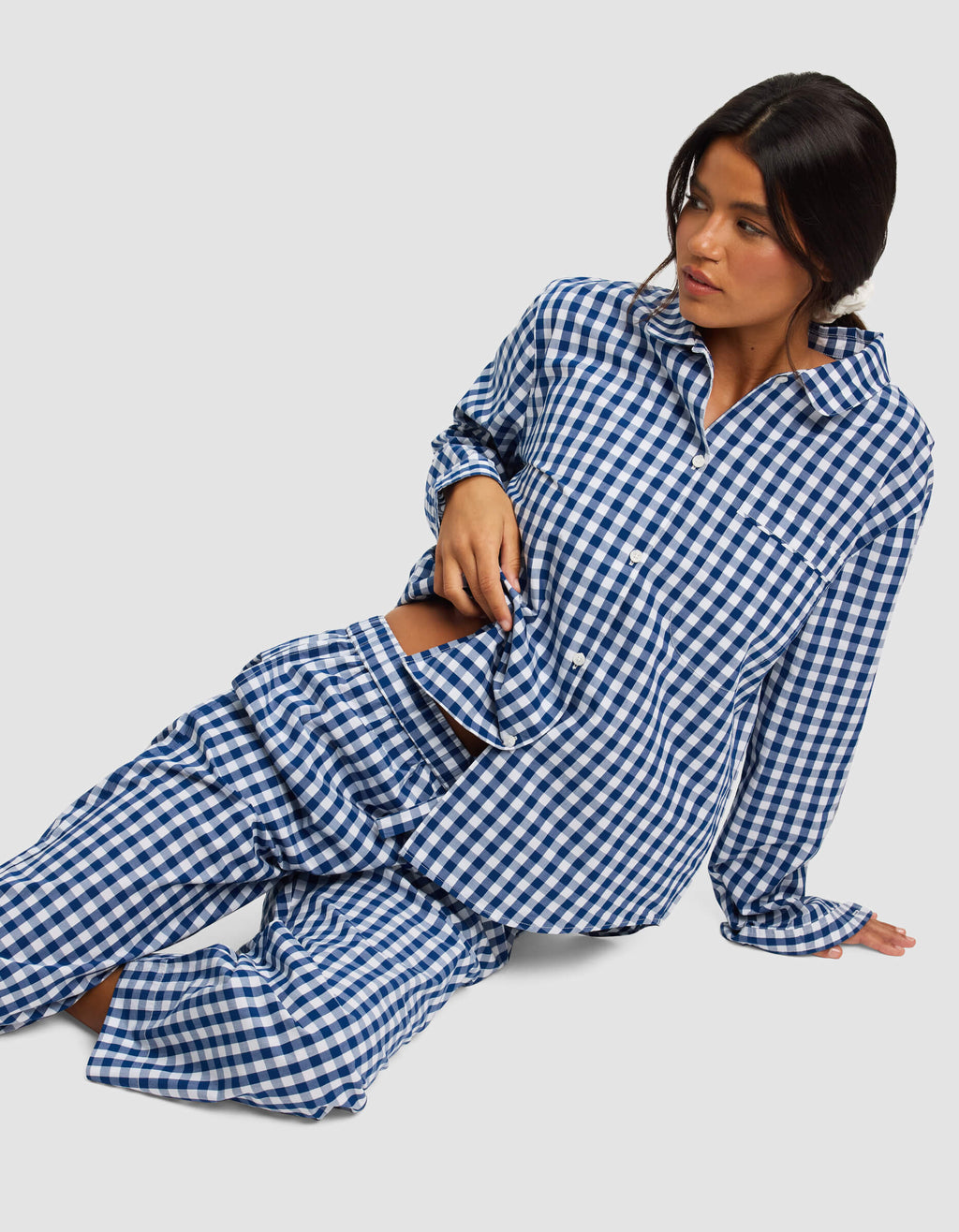 A woman with long dark hair wears the Cozy Earth Women's Sutton Long Sleeve Pajama Top in blue and white gingham, sitting on the floor and leaning to one side, looking off to her left against a plain, light background.|Color:Pacific Gingham