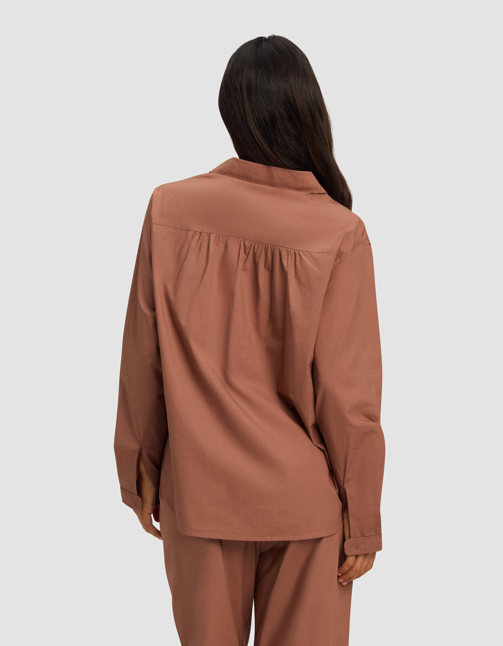 A woman with long dark hair is seen from behind wearing the Cozy Earth Women's Sutton Long Sleeve Pajama Top in rust against a plain light background.|Color:Saddle