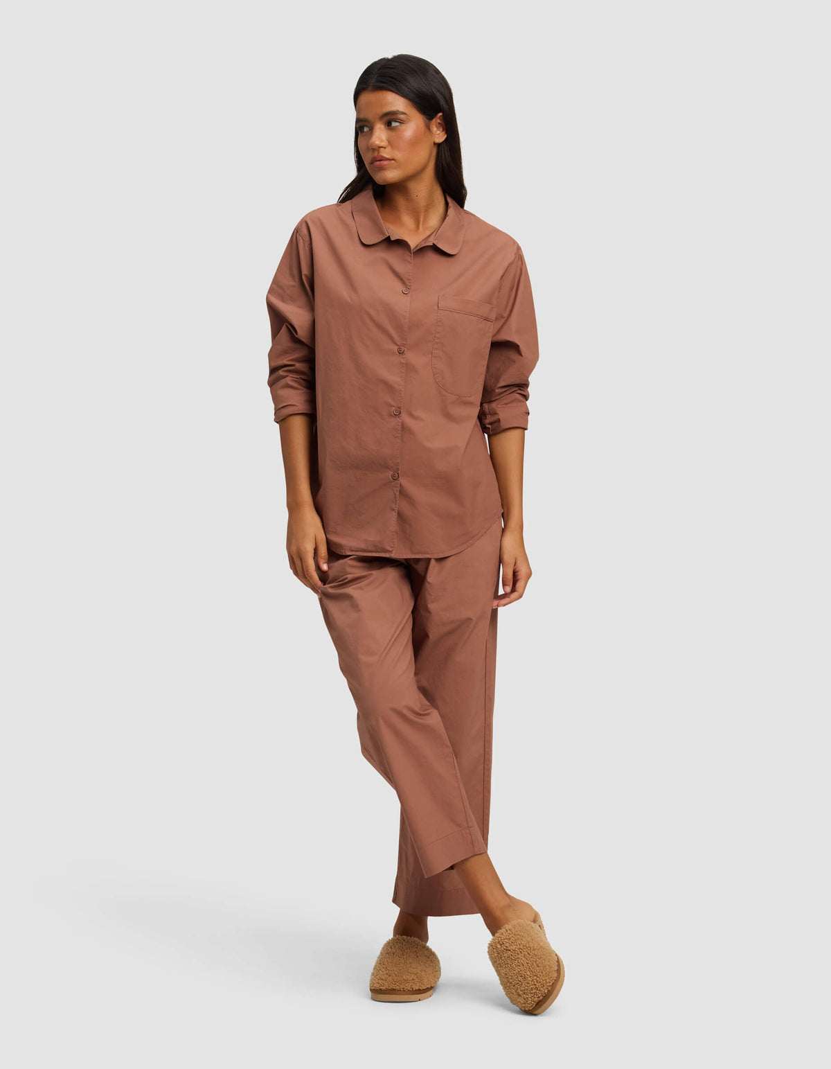 A person with long dark hair and a relaxed posture stands against a plain background, wearing the Cozy Earth Women's Sutton Long Sleeve Pajama Top in brown, matching pants, and beige fuzzy slippers.|Color:Saddle