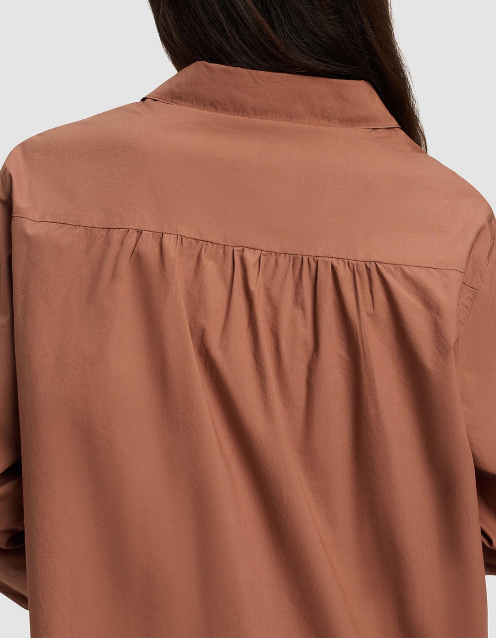 A woman with long brown hair is seen from behind wearing the Cozy Earth Women's Sutton Long Sleeve Pajama Top in rust, featuring loose fit and gathered fabric detail below the yoke.|Color:Saddle