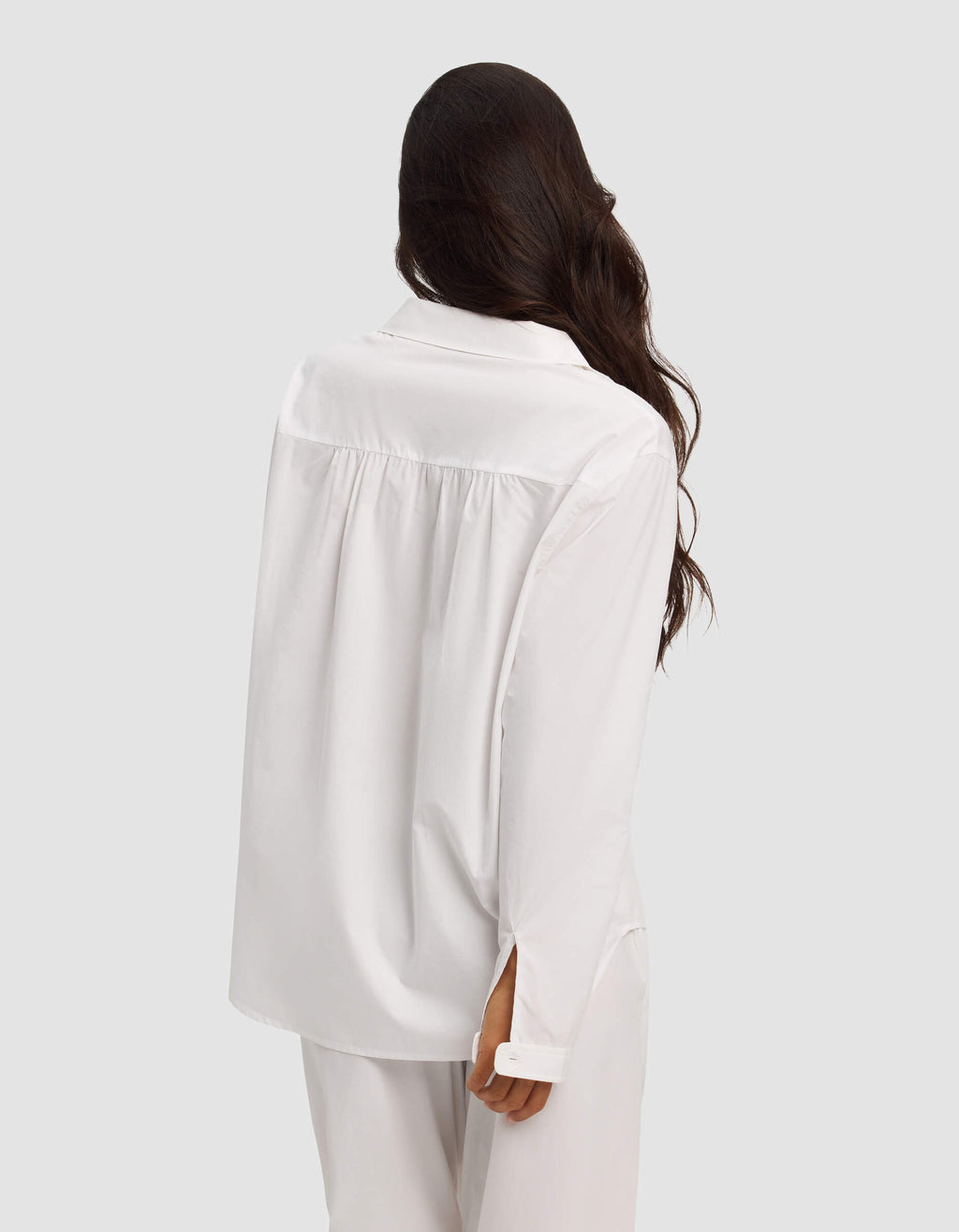 A woman with long, wavy dark hair faces away, wearing the Cozy Earth Women's Sutton Long Sleeve Pajama Top in white against a plain light gray background.|Color:White