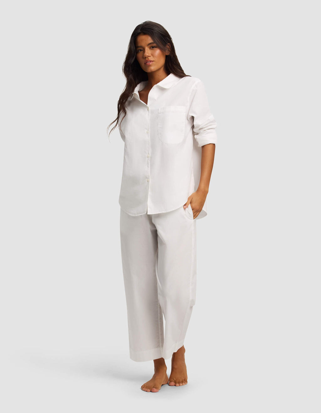A woman with long dark hair stands barefoot against a plain background, wearing the Cozy Earth Women's Sutton Long Sleeve Pajama Top in white and matching wide-leg pants, styled with rolled sleeves and a relaxed, neutral expression.|Color:White