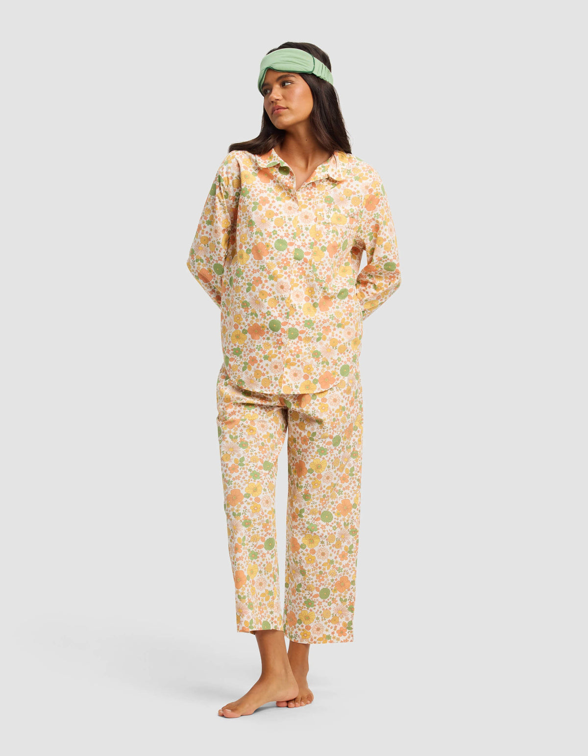 A woman stands barefoot against a plain light gray background, wearing the Cozy Earth Women’s Sutton Long Sleeve Pajama Top in a loose, floral pastel design and a green sleep mask.|Color:Wildflower