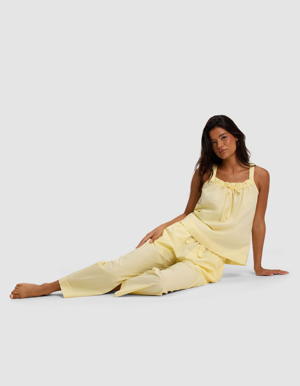 A woman with long dark hair sits on the floor against a light grey background, wearing Cozy Earth’s Women's Sutton Cropped Pajama Pant—a sleeveless yellow pajama set with loose-fitting pants and a ruffled neckline.|Color:Meringue