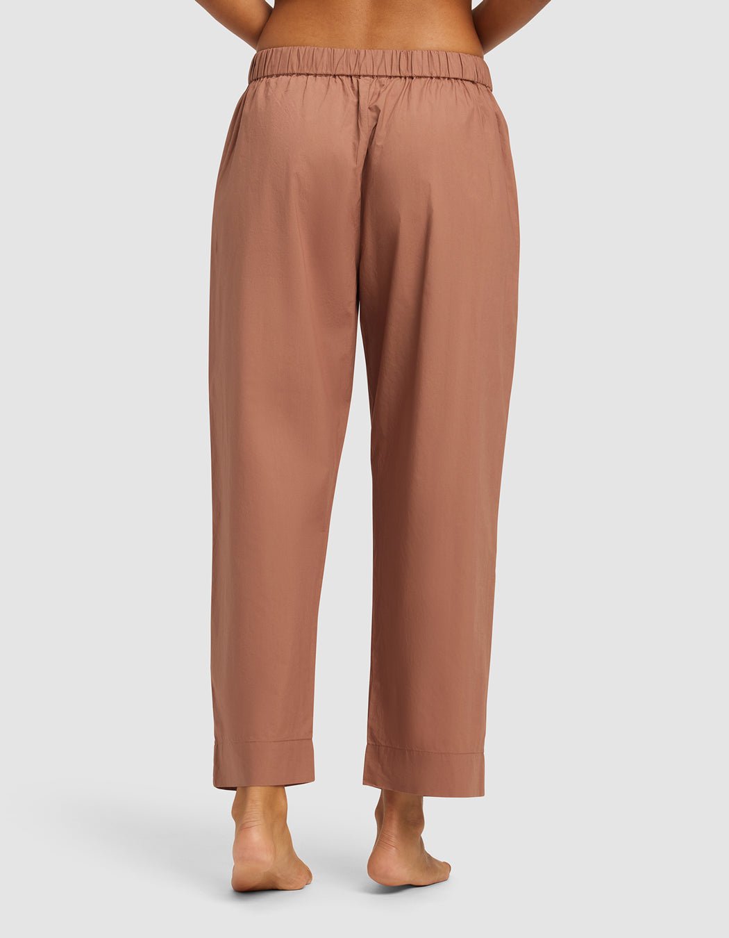 A person stands barefoot, shown from the waist down, wearing Cozy Earth's Women's Sutton Cropped Pajama Pant in brownish-pink with an elastic waistband and cuffed hems. The background is plain and light-colored.|Color:Saddle