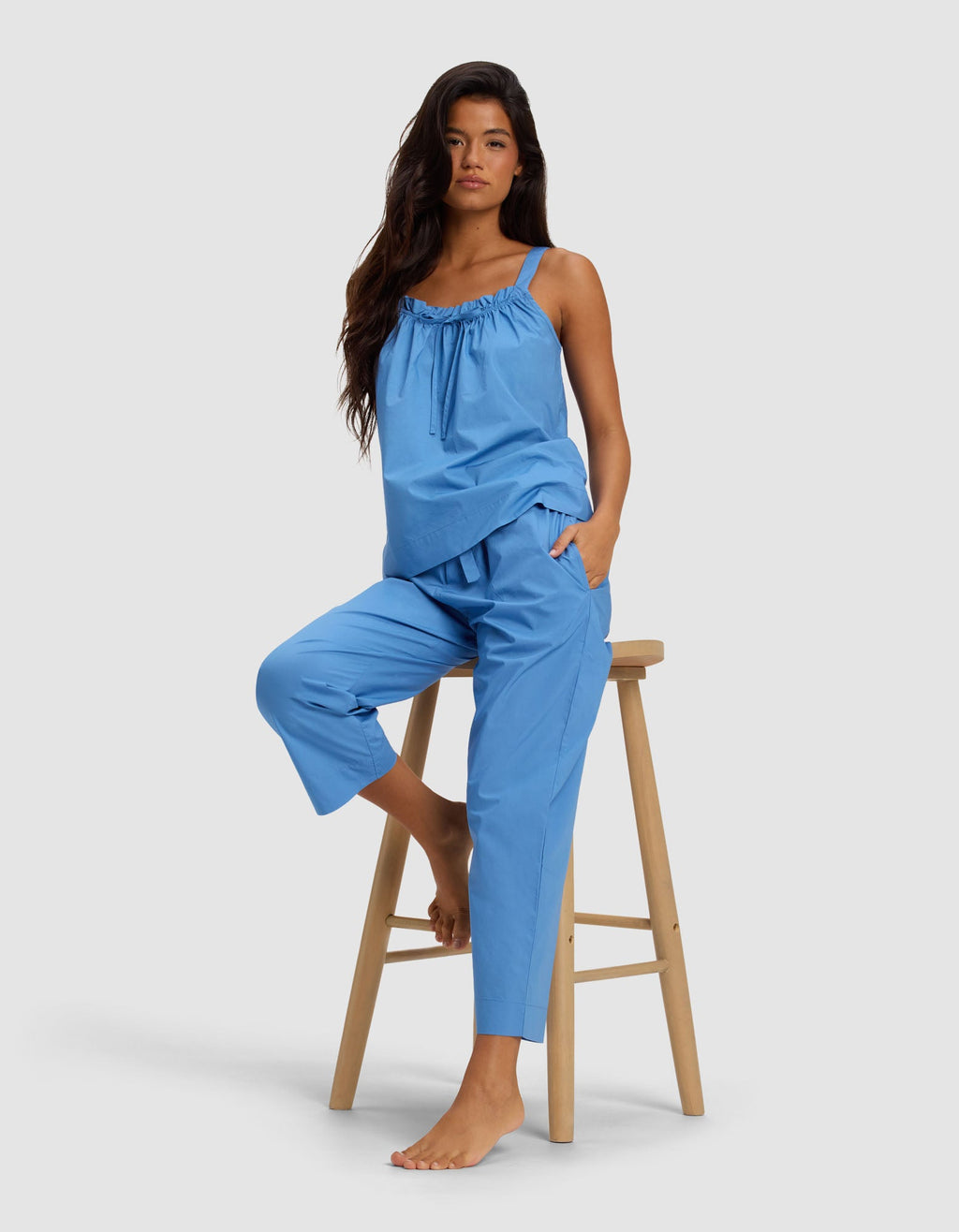 A woman with long dark hair models Cozy Earth's Women's Sutton Cropped Pajama Pant in blue, paired with a matching sleeveless top, as she sits barefoot on a wooden stool against a plain light background.|Color:Sailor
