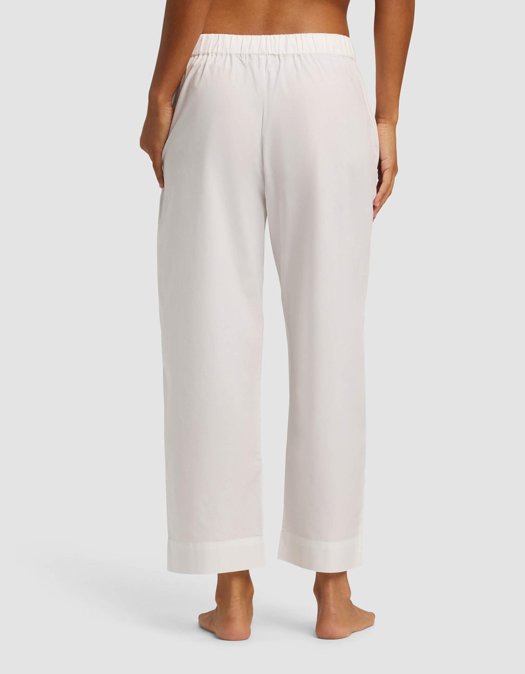 A woman stands barefoot, facing away from the camera, wearing Cozy Earth's Women's Sutton Cropped Pajama Pant—loose-fitting, light-colored with an elastic waistband and pockets—against a plain light background.|Color:White