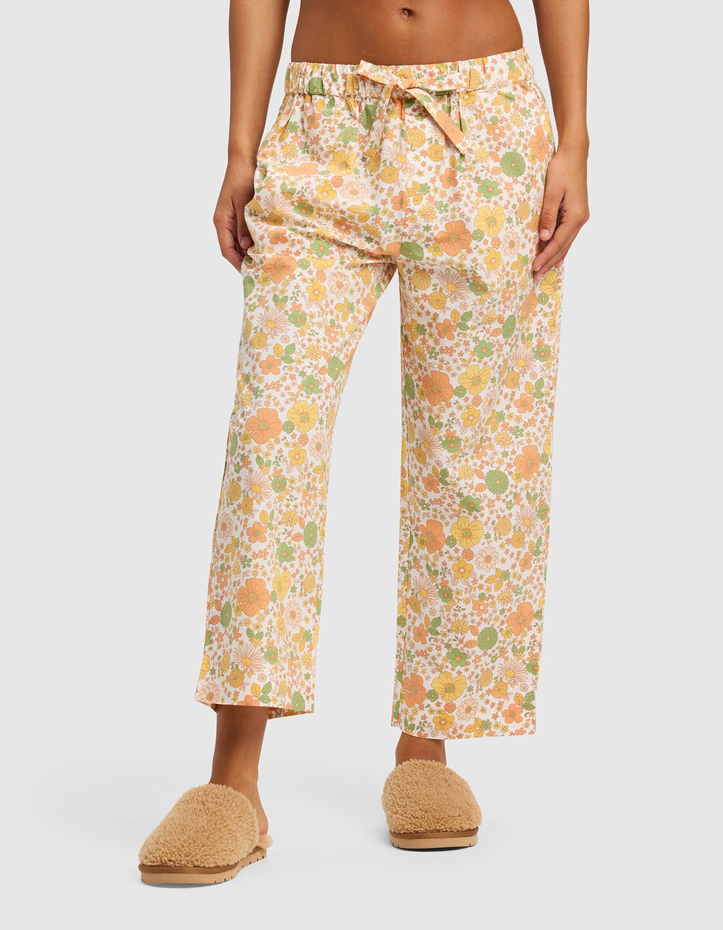A person wears Cozy Earth's Women's Sutton Cropped Pajama Pant with a colorful floral pattern and tan fuzzy slippers, standing against a light gray background.|Color:Wildflower