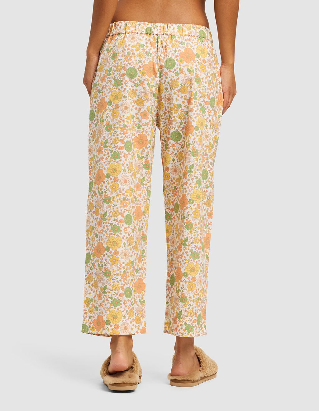 A person stands barefoot with their back to the camera, wearing Cozy Earth's Women's Sutton Cropped Pajama Pant in a loose floral print, paired with fuzzy beige slippers, against a plain background.|Color:Wildflower