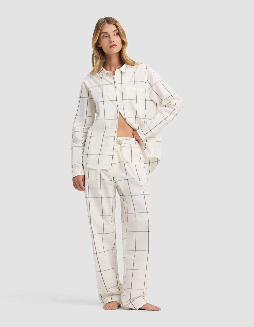 A woman stands barefoot against a light gray background, wearing Cozy Earth's Women's Sutton Pajama Pant in white with a black grid pattern and a loose button-up shirt partly tucked in, revealing her midriff.|Color:Bone Windowpane