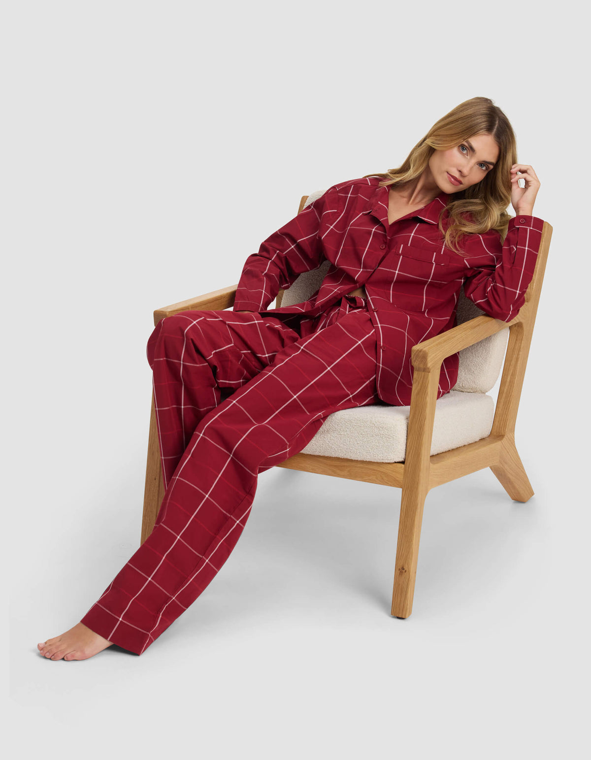 A woman sits barefoot on a wooden chair, wearing the Cozy Earth Women's Sutton Pajama Pant in red with white grid lines. She leans back, one arm on the armrest, and tilts her head slightly to the side.|Color:Garnet Windowpane