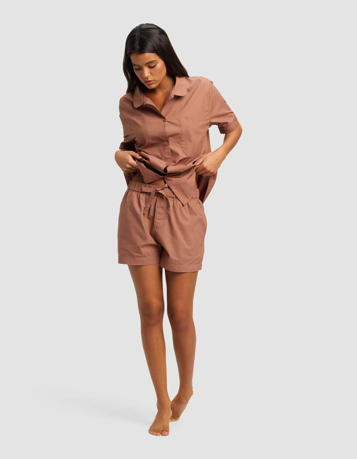A woman wearing Cozy Earth's Women's Sutton Pajama Short with a matching loose, short-sleeved, button-up shirt stands barefoot against a plain light background, looking down and adjusting her shirt.|Color:Saddle