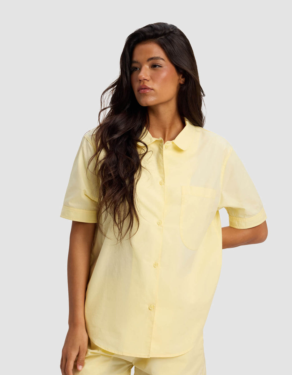 A woman with long, wavy dark hair models the Cozy Earth Women's Sutton Short Sleeve & Cropped Pant Pajama Set in light yellow, standing against a plain light gray background and looking to her left.|Color:Meringue