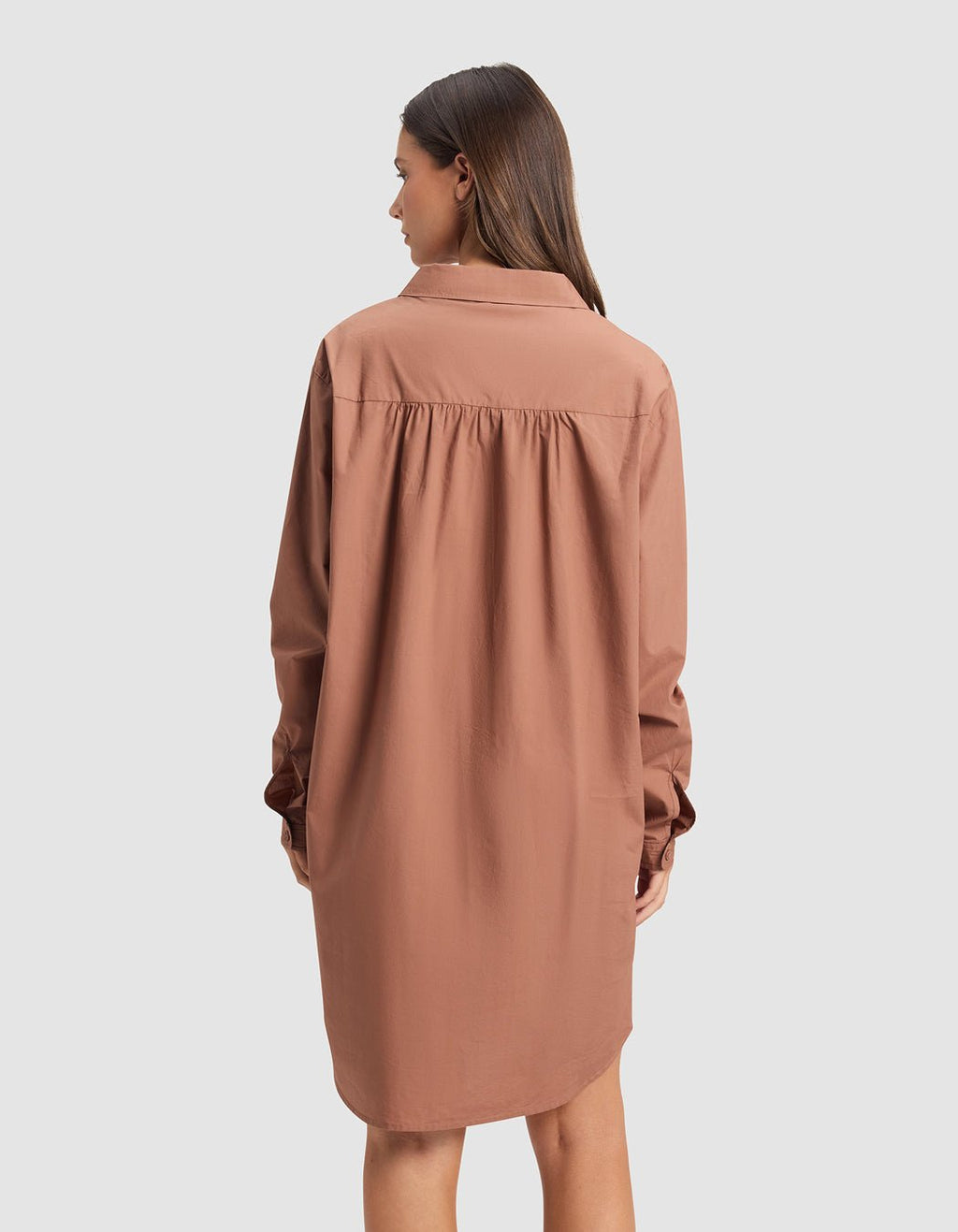 A woman faces away from the camera, wearing Cozy Earth's Women's Sutton Sleep Dress—a loose, long-sleeved, knee-length brown shirt dress with a collar—against a plain light gray background.|Color:Saddle