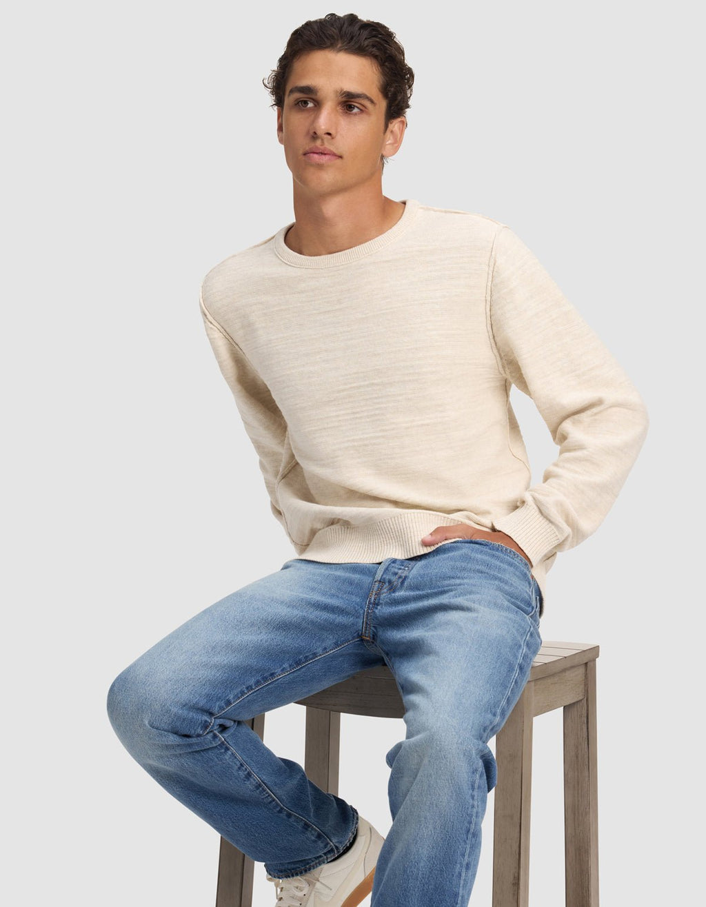 A young man with short curly hair wears the Cozy Earth Men's Tate Crewneck and blue jeans while seated on a wooden stool against a plain light gray background, gazing slightly to the side.|Color:Alabaster