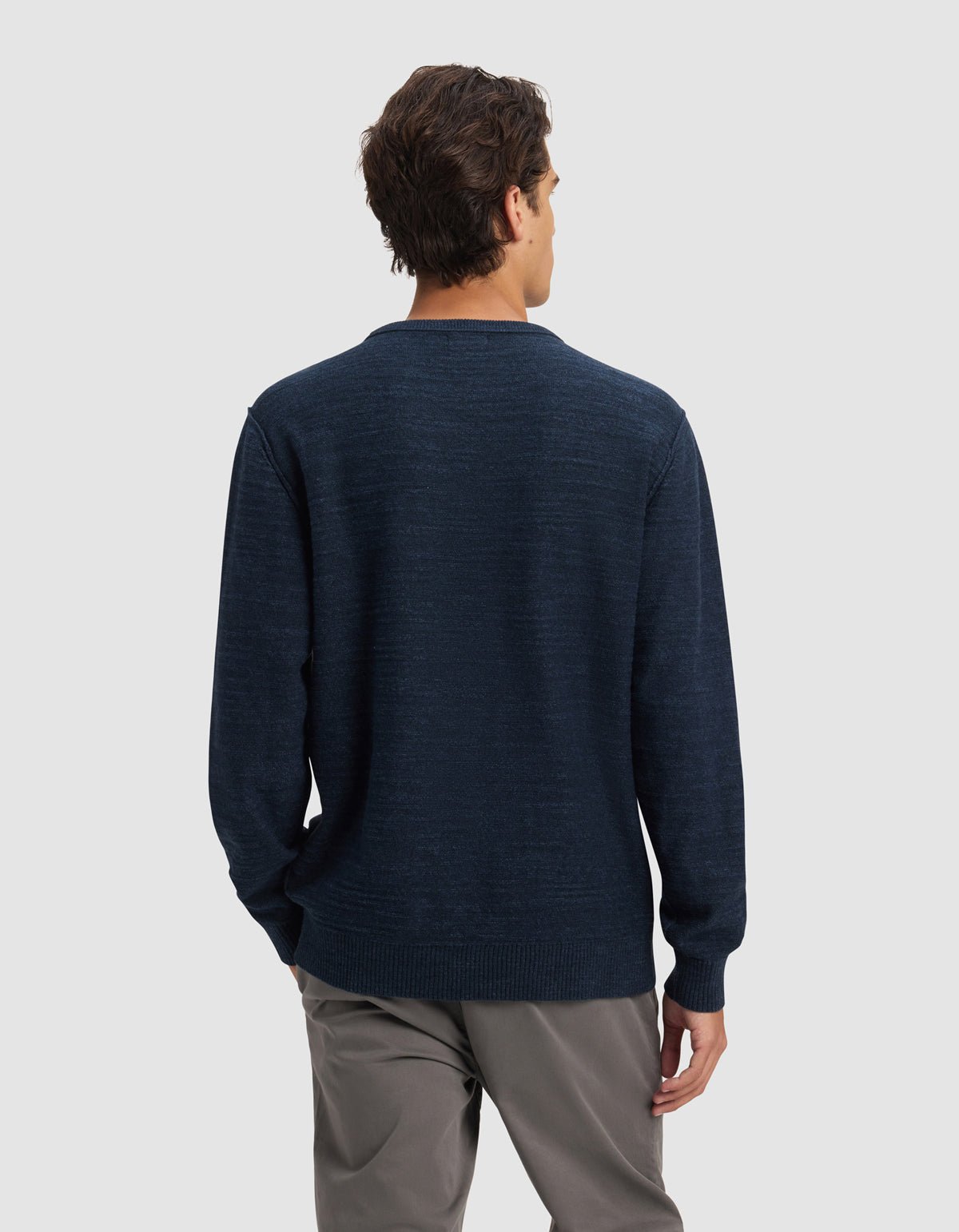 A man stands facing away from the camera, wearing Cozy Earth's Men's Tate Crewneck in dark blue and gray pants against a plain light gray background.|Color:Eclipse