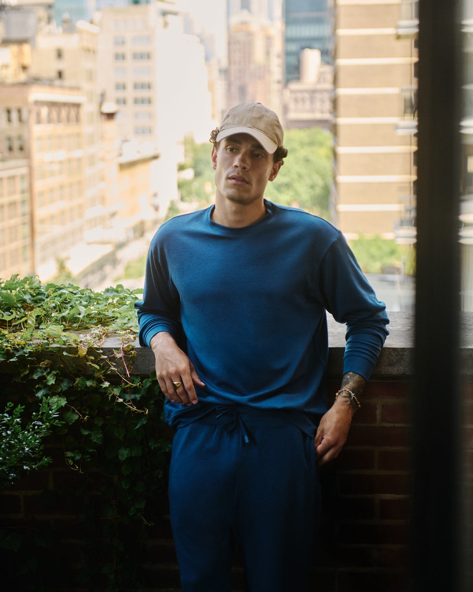 A man wearing a blue Cozy Earth Versa crewneck and matching blue Versa Joggers stands on a balcony with a city skyline background.|Color:Obsidian