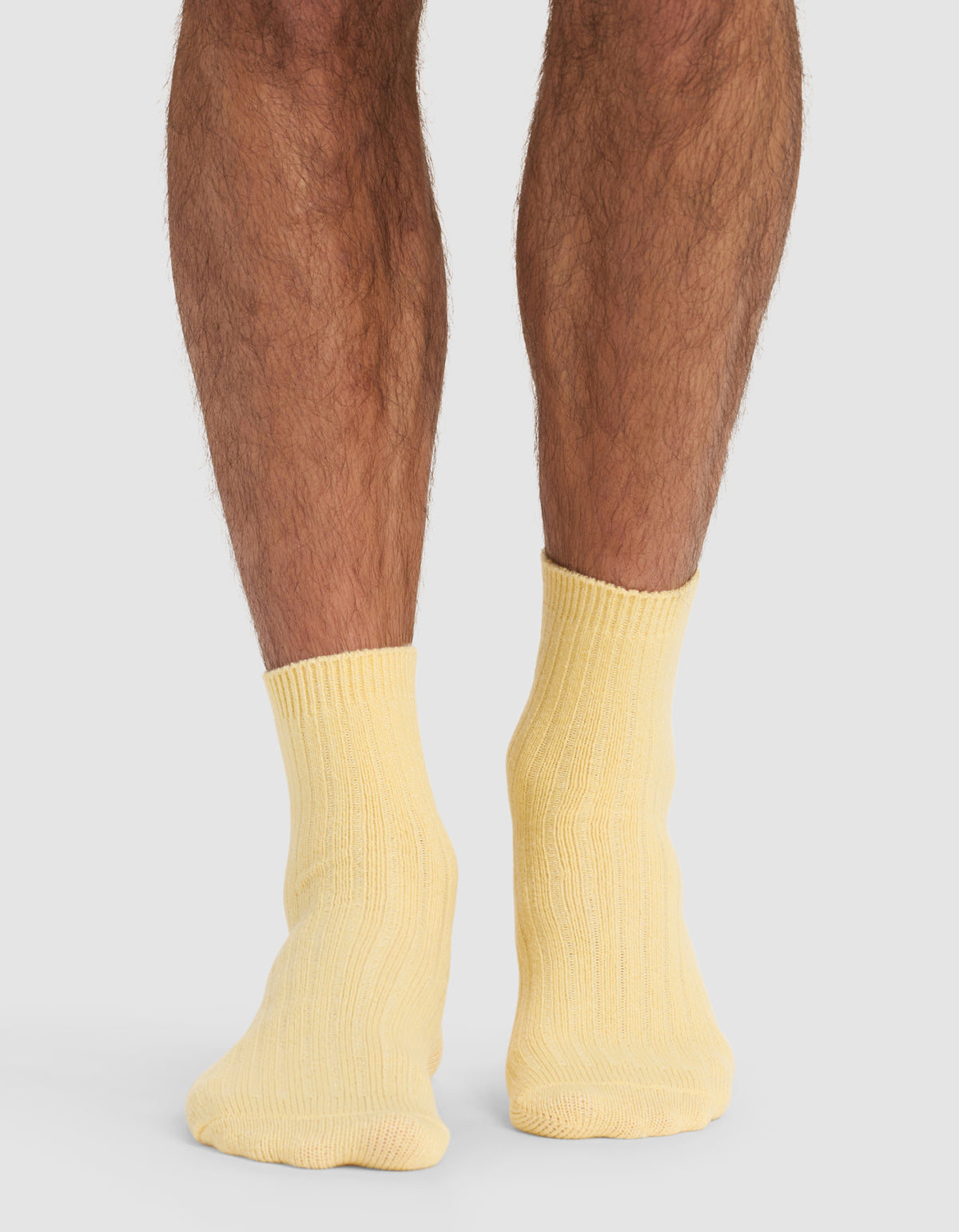 A person stands on a light surface, shown from the calves down, wearing Cozy Earth's Plush Lounge Quarter Socks in pale yellow. Their feet are close together, set against a neutral gray background.|Color:Daisy