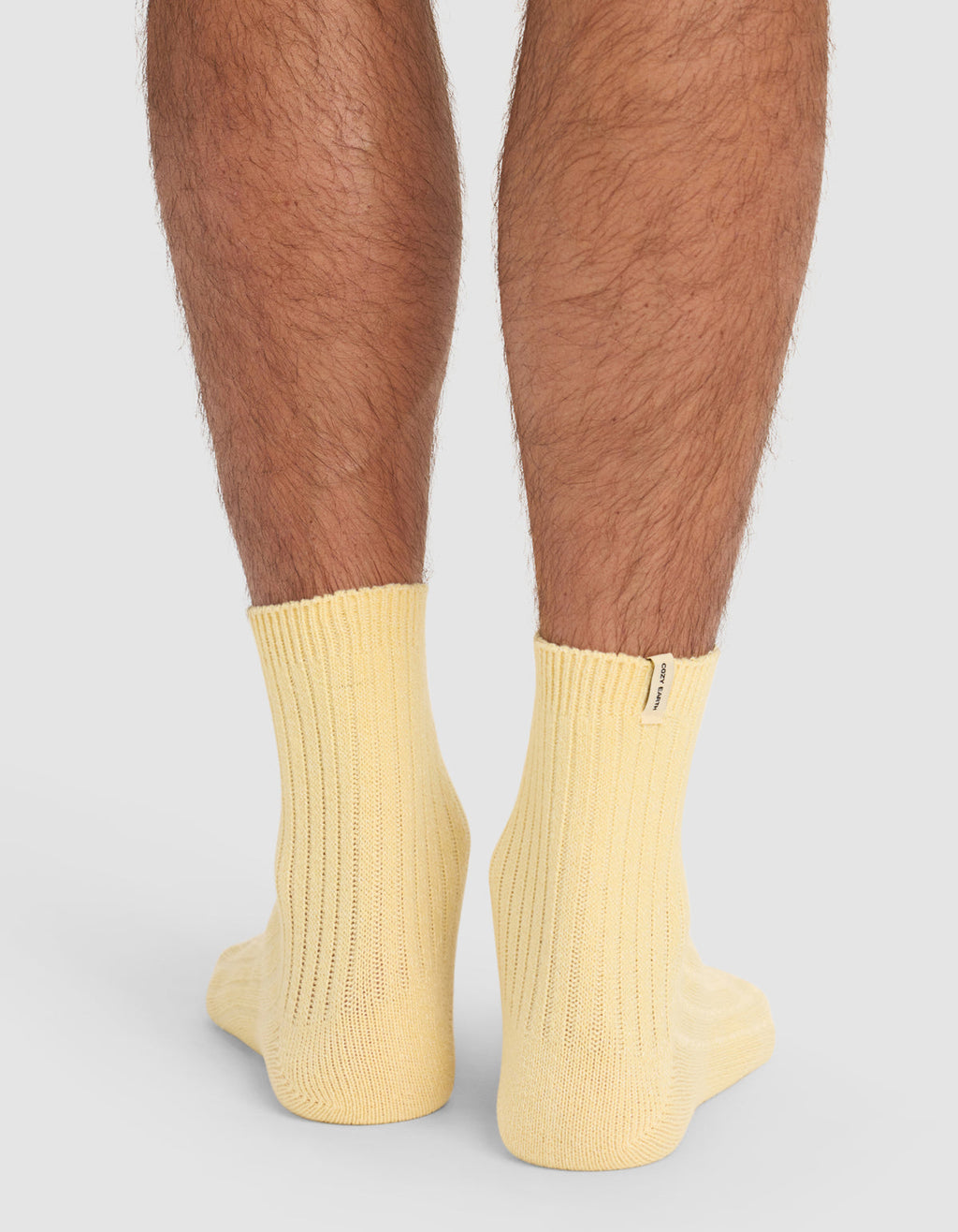 The back view of a person's lower legs wearing Cozy Earth's Plush Lounge Quarter Socks in pale yellow ribbed fabric, standing on a white surface.|Color:Daisy
