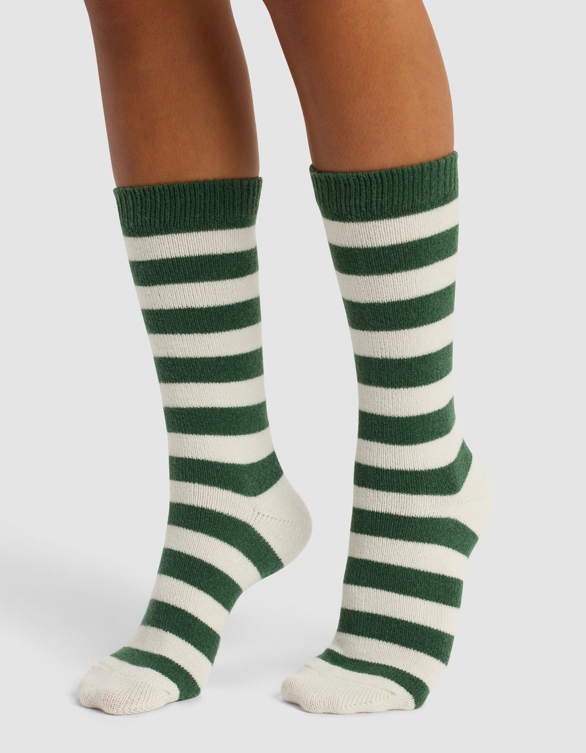 A pair of feet wear Cozy Earth's The Striped Plush Lounge Sock—knee-high, with horizontal dark green and white stripes—standing on a white background.|Color:Celadon Stripe