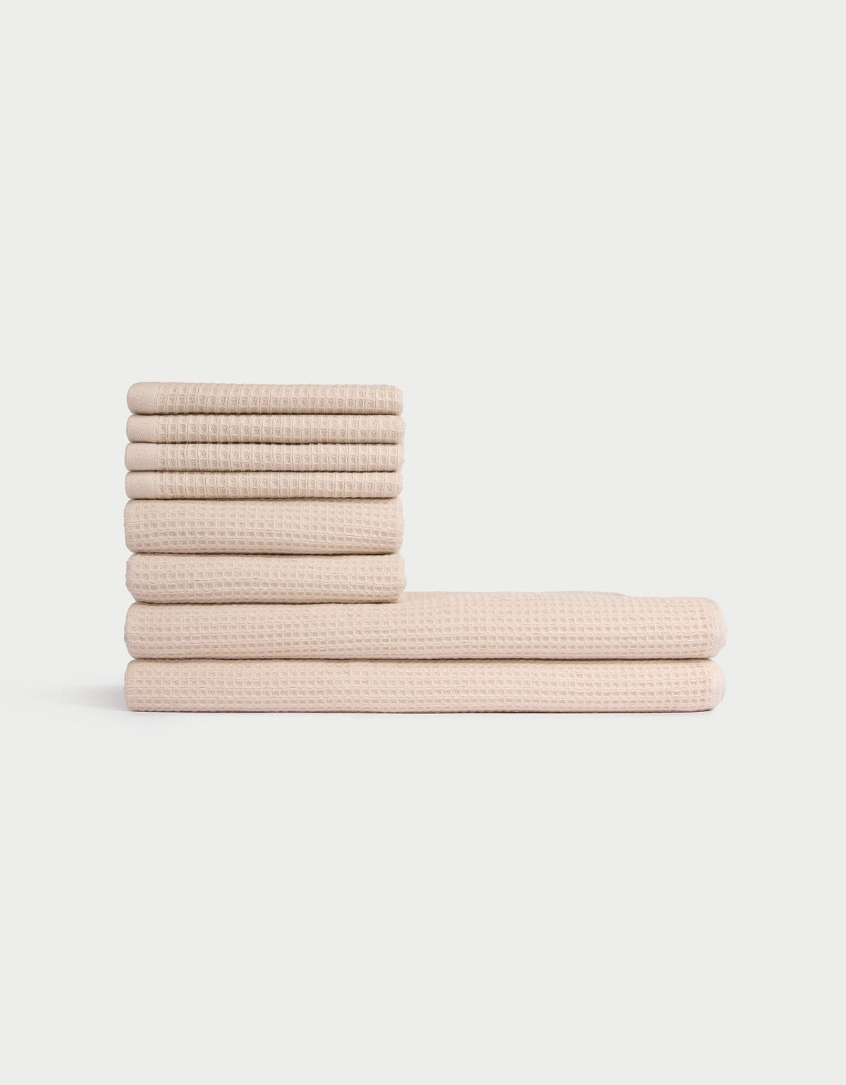 The Cozy Earth Waffle Bath Towel Set features eight neatly folded beige towels with a textured waffle pattern, stacked in two piles against a plain white background. |Color:Birch