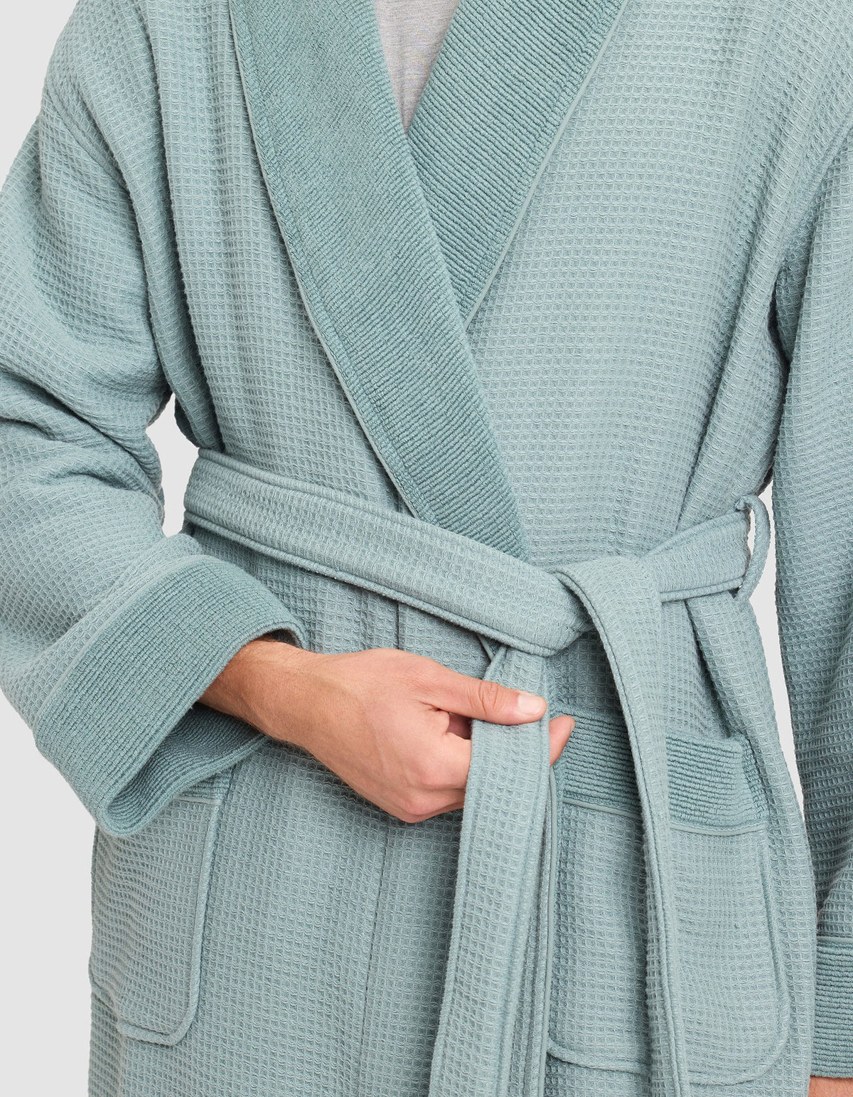 A person is tying the belt of a Cozy Earth Waffle Bath Robe in light blue, holding the belt with their left hand and adjusting it with their right. The textured robe is visible from the shoulders down.  |Color:Brook