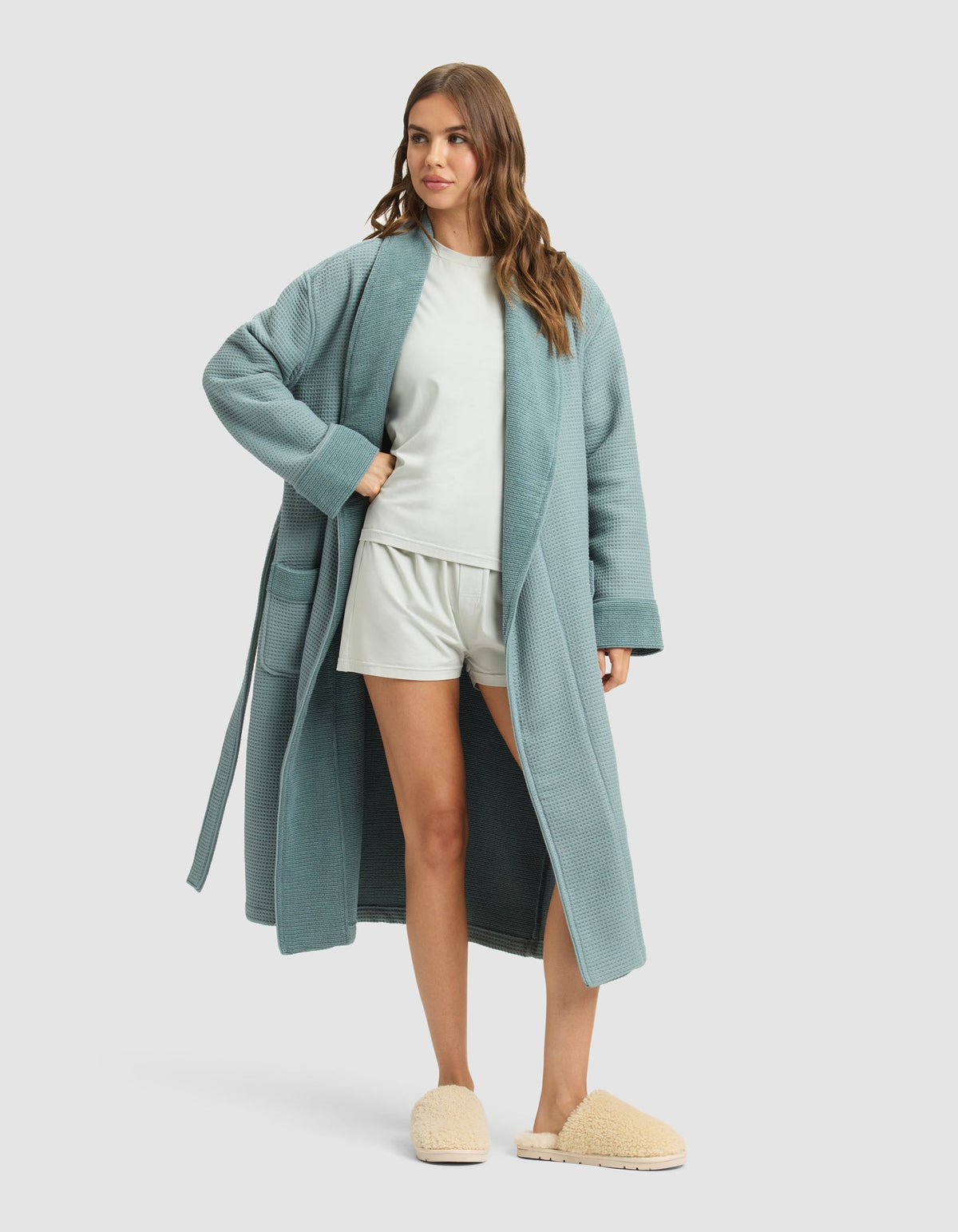 A woman with wavy brown hair models the Cozy Earth Waffle Bath Robe in teal over a white T-shirt and shorts with beige slippers, posing against a plain light gray background. |Color:Brook