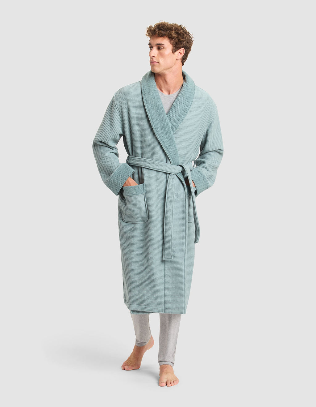 A man stands barefoot, looking to the side, wearing Cozy Earth’s Waffle Bath Robe in light blue with a belt and two front pockets over light gray pants.  |Color:Brook