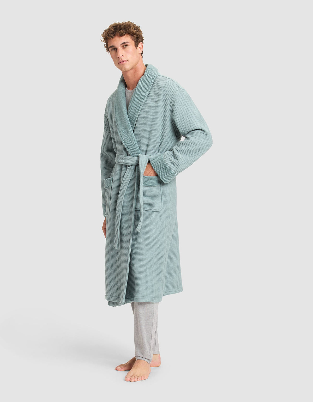 A man stands barefoot in Cozy Earth's Waffle Bath Robe, a light blue-green, waffle-knit robe with a belt and two front pockets, paired with light gray pajama pants, against a plain light background.  |Color:Brook