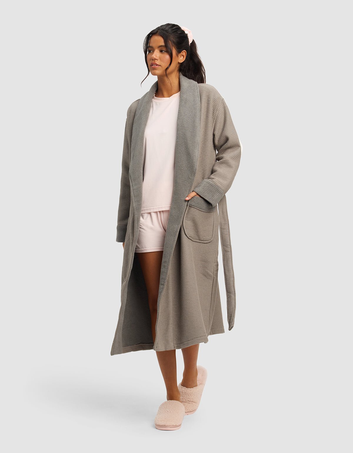 A woman stands against a plain background, wearing the Cozy Earth Waffle Bath Robe in gray over a light pink pajama set and pink fuzzy slippers, with one hand in her pocket and a relaxed expression.|Color:Charcoal
