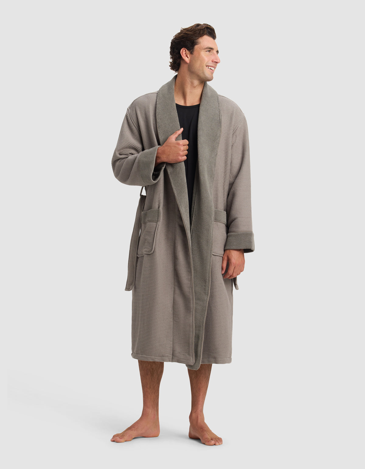 A man stands barefoot, smiling and looking to the side, wearing Cozy Earth's Waffle Bath Robe in gray over a black shirt. The robe features large pockets, a belt, and is set against a plain light gray background.|Color:Charcoal
