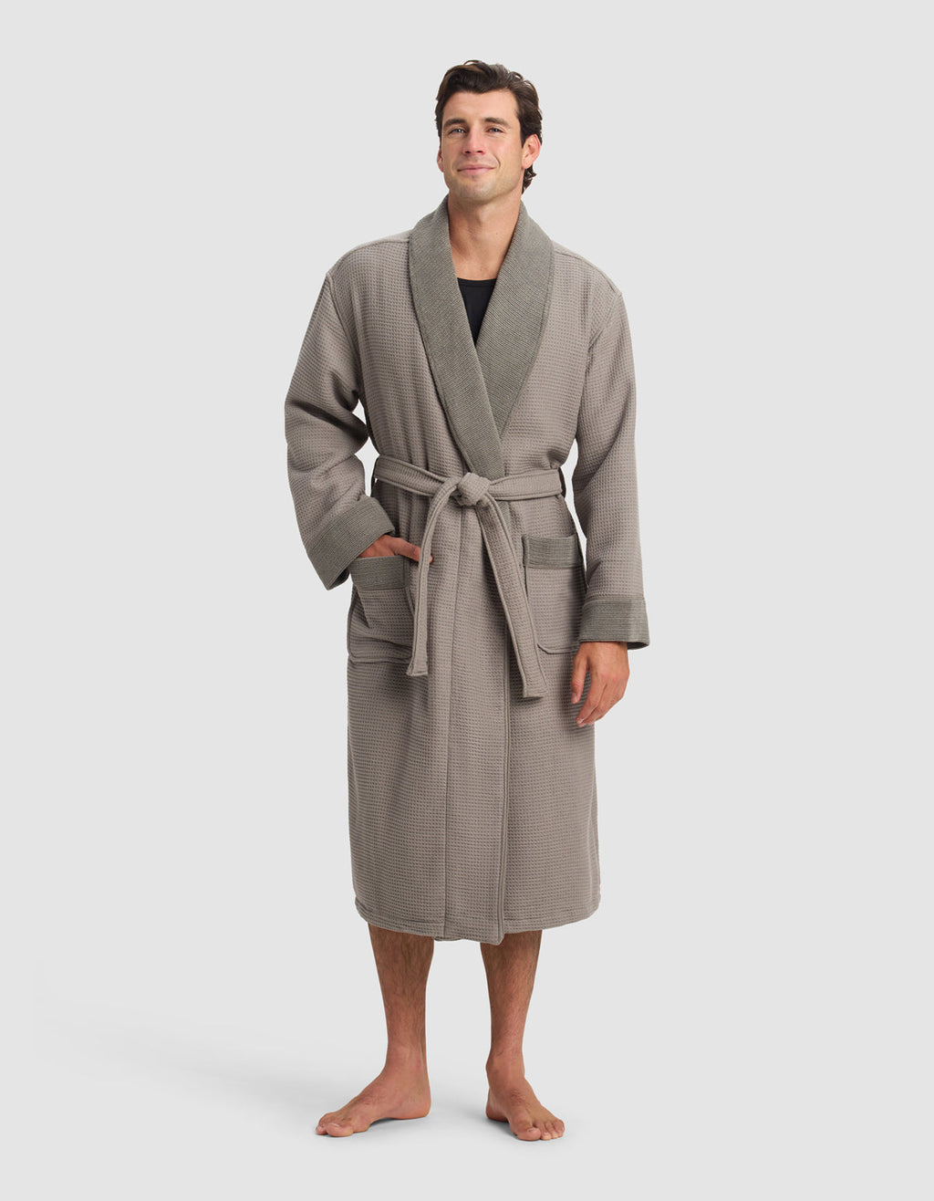 A man stands barefoot in a Cozy Earth Waffle Bath Robe—light brown with a darker shawl collar, front pockets, and a tied belt—worn over a black shirt. The background is plain and light-colored.|Color:Charcoal