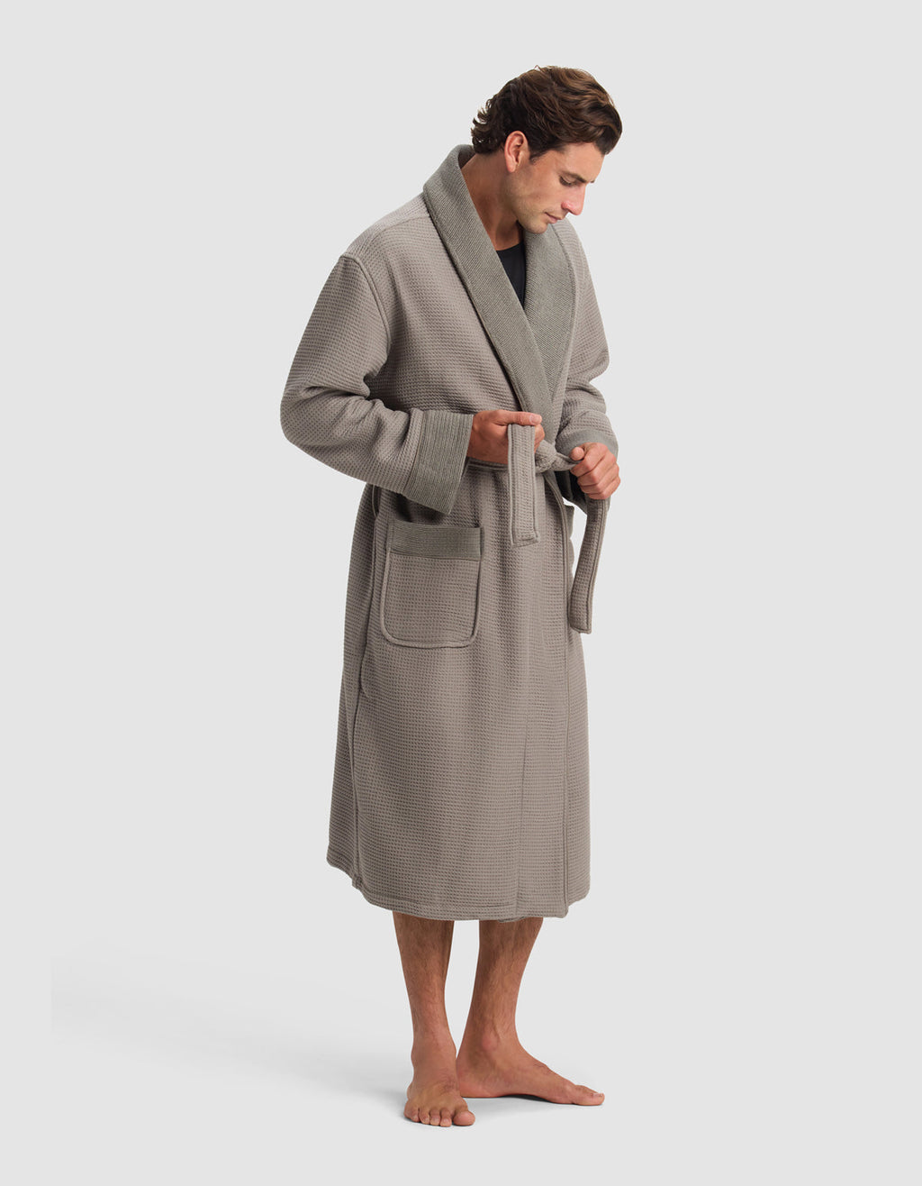 A man barefoot in a Cozy Earth Waffle Bath Robe stands against a plain light background, looking down as he ties the robe's belt at his waist.|Color:Charcoal
