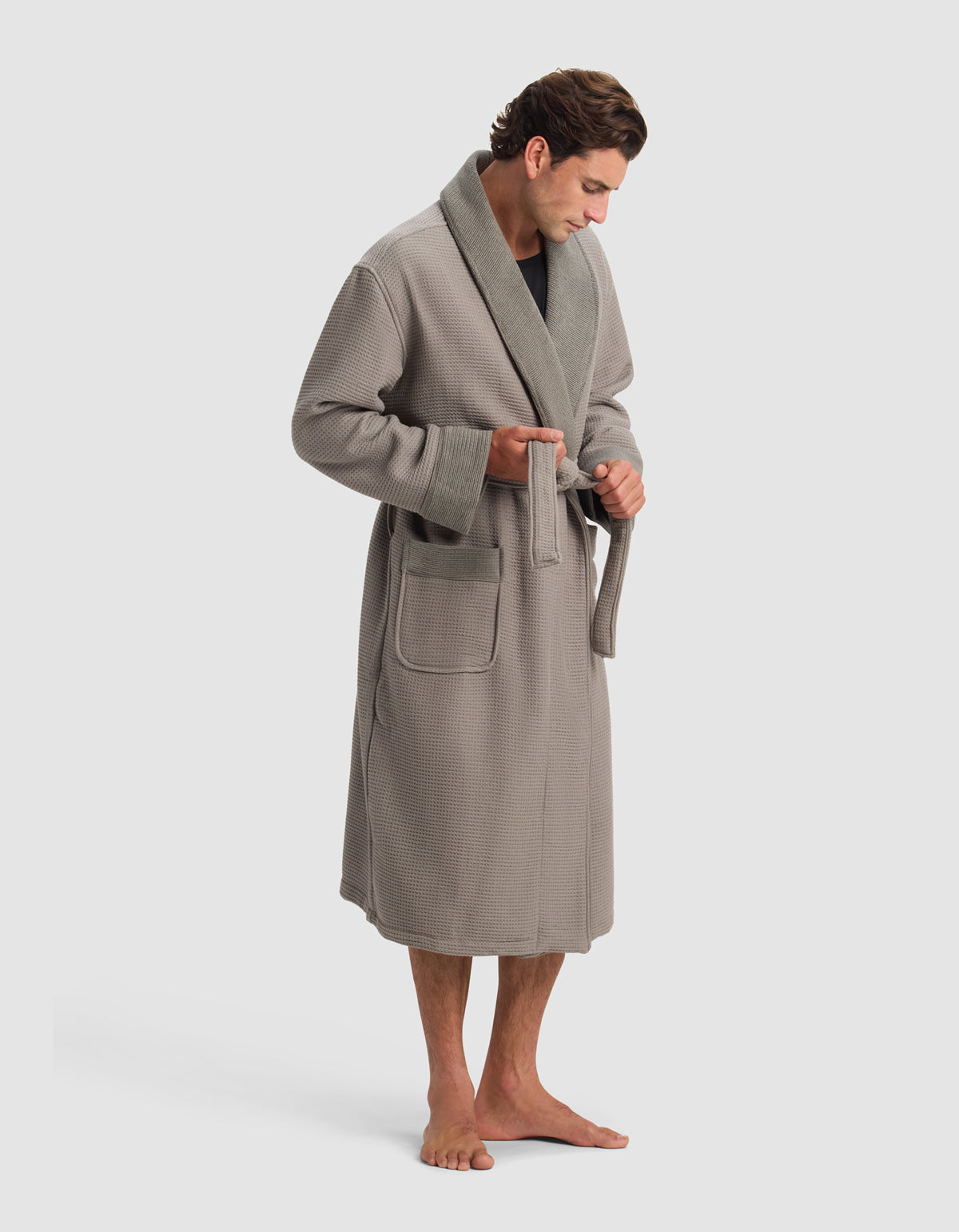 A man barefoot in a Cozy Earth Waffle Bath Robe stands against a plain light background, looking down as he ties the robe's belt at his waist.|Color:Charcoal