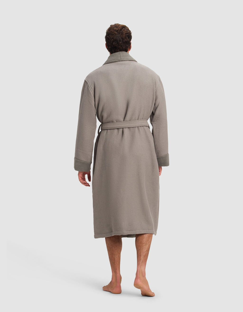 A man stands barefoot, facing away from the camera, wearing the Cozy Earth Waffle Bath Robe—a long, light gray, textured robe with a belted waist.|Color:Charcoal