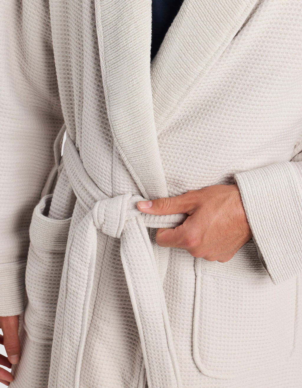 A person is shown tying the belt of a Cozy Earth Waffle Bath Robe at the waist. The light-colored, textured robe features large front pockets, with only the wearer’s torso and hands visible.|Color:Light Grey
